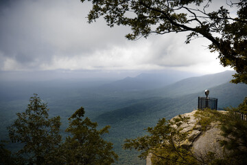 Dramatic clifftop view of a vast, misty green valley under a heavy, moody sky. A coin-operated telescope on the edge provides a travel and adventure focus. Caesars Head State Park