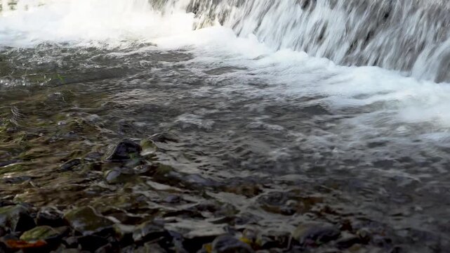 A low-angle shot showing a waterfall or small rapids falling into a river below. The waterfall creates white foam, while the foreground water ripples and shimmers over small rocks on the riverbed.