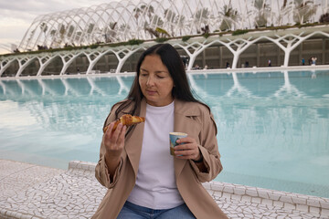 Woman holding coffee and pastry near water. Relaxed lifestyle moment.