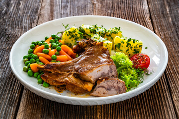 Baked veal cutlet with bone, boiled potatoes, mini carrots and green peas on wooden table