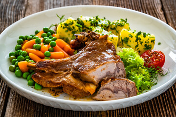 Baked veal cutlet with bone, boiled potatoes, mini carrots and green peas on wooden table