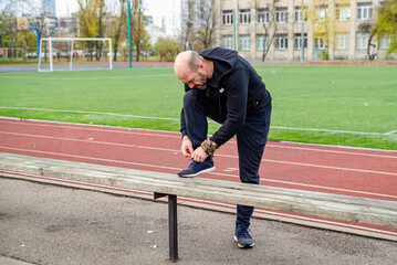 A focused man ties his shoelaces on a wooden bench at an outdoor athletic track, preparing for a run or workout. Emphasizes readiness, an active lifestyle, and sports fitness.