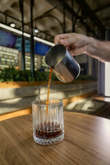 Barista pouring hot coffee into a glass on a wooden table in a modern cafe