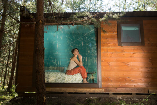 A tired sad middle aged woman sits at the window of a country house in the forest. work burnout. solo retreat