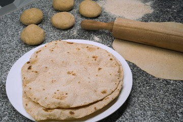Making roti the unleavened flatbread. Close-up of several cooked roti on a white plate. Nearby are flour, a rolling pin, and balls of dough on a gray granite kitchen surface. Backing homemade bread.