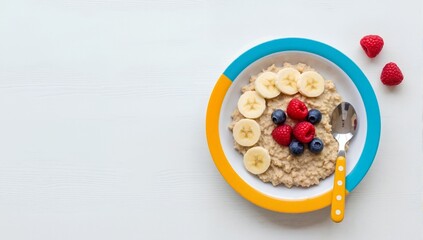 Oatmeal porridge with banana, blueberries and raspberries in a colorful bowl. Healthy breakfast for kids on white wooden background. Flat lay concept with copy space