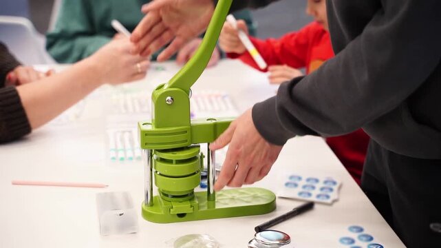 Hands on a device make badge on clothing with children's drawings on a white table where children and adults are sitting