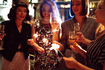 Happy women in stylish outfits are having fun in cozy room, clinking glasses and lighting sparklers. Four girlfriends are enjoying holiday with glasses and sparklers. The concept is cozy and fun.