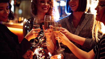 Happy women in stylish outfits are having fun in cozy room, clinking glasses and lighting...