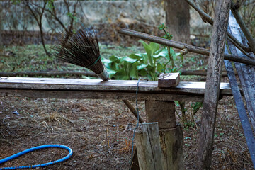 simple, outdoor utility shelf or bench constructed from rough wooden planks. A short, natural-fiber broom (hand broom) and a small wooden block rest on the shelf. Beneath, a blue garden hose lies coil