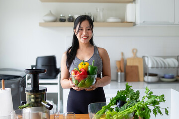 A young woman holds a cup containing various vegetables, preparing to make vegetable juice in the kitchen.