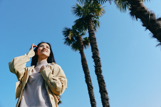 Sunlit scene featuring a woman looking up toward a clear blue sky beside tall palm trees, wearing a light jacket, conveying freedom, outdoor travel, and carefree mood. - Powered by Adobe
