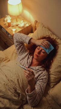 Vertical video of a sick woman lying in bed at night with a fever. Female checking her temperature with a thermometer while wearing a cooling patch on her forehead. Flu and illness concept