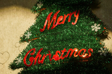 close-up shot of a tinsel Christmas tree decoration featuring bright, red glitter script spelling "Merry Christmas," capturing the texture and sparkle of homemade or festive holiday signage.