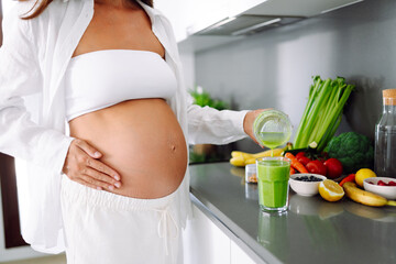 Pregnant woman stands in modern kitchen, pouring green smoothie into glass. Expectant mother stands near countertop with fresh vegetables and fruit in cozy space. Concept of health, and beverages.