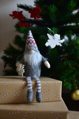 Cozy Christmas scene with a small Nordic gnome sitting on wrapped gifts under a decorated tree. Kraft paper boxes, soft handmade toy and natural holiday decor create a warm festive atmosphere at home.