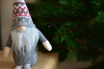 Cozy Christmas scene with a small Nordic gnome sitting on wrapped gifts under a decorated tree. Kraft paper boxes, soft handmade toy and natural holiday decor create a warm festive atmosphere at home.