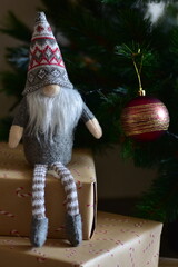 Cozy Christmas scene with a small Nordic gnome sitting on wrapped gifts under a decorated tree. Kraft paper boxes, soft handmade toy and natural holiday decor create a warm festive atmosphere at home.