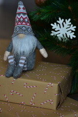 Cozy Christmas scene with a small Nordic gnome sitting on wrapped gifts under a decorated tree. Kraft paper boxes, soft handmade toy and natural holiday decor create a warm festive atmosphere at home.