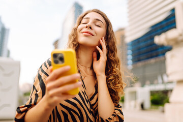 A portrait of a young woman smiling with a phone, standing against the backdrop of a metropolitan...
