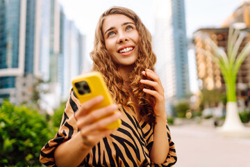 A portrait of a young woman smiling with a phone, standing against the backdrop of a metropolitan...