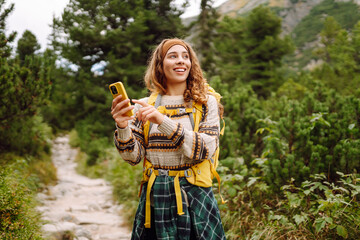 Female hiker with yellow backpack and phone walks along hiking trail on sunny day. Woman enjoys mountain scenery while hiking, texts or blogs on phone. Concept of adventure, technology, blogging.