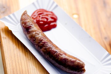 Grilled sausage on a white paper plate, accompanied by a dollop of red ketchup, resting on a wooden table, showcasing delicious street food presentation and culinary delight