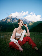 Obraz premium Outdoor landscape scene with a model seated in a grassy field, mountains in the distance, wearing a white crop top and red pants, a towel over shoulders, capturing a calm, nature infused moment.