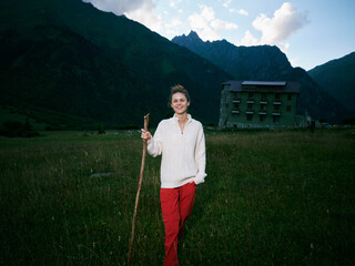 Obraz premium Woman standing in a grassy field with a wooden staff, set against distant mountains and a rustic building, outdoor adventure scene, calm atmosphere, hiking gear and nature backdrop