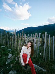 Obraz premium Woman sits by a weathered wooden fence in a mountainous landscape, wearing a light sweater and red pants, exploring nature and countryside ambience under a blue sky with scattered clouds.