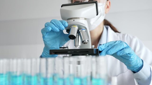 Female scientist wearing protective equipment checking microscope, analyzing blue test tubes in modern laboratory research setting. Medicine, healthcare and science concept