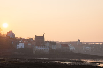 Golden sunset light bathes the historic seafront buildings of South Queensferry beside the Firth of Forth, with a hazy view toward the Forth Road Bridge on a calm misty evening in Scotland.