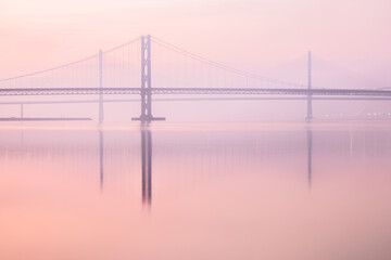 The Forth Road Bridge at dusk through soft pink mist over the Firth of Forth, its elegant suspension towers and reflections creating a calm, minimalist scene in South Queensferry, Scotland.