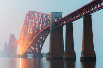 The iconic Forth Bridge spans the calm waters of the Firth of Forth near South Queensferry, Scotland, its red cantilever structure glowing through the  mist during blue hour.