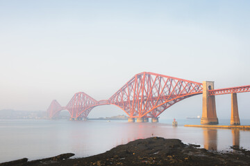 The iconic Forth Bridge spans the calm waters of the Firth of Forth near South Queensferry, Scotland, its red cantilever structure glowing through the morning mist under a pale blue sky.