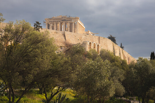 The Parthenon atop the Acropolis of Athens stands bathed in golden light against dramatic clouds, surrounded by olive trees and ancient stone walls undergoing restoration in Greece.