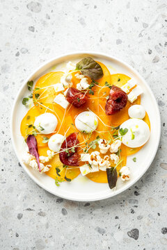 Vibrant, elegant plated appetizer of yellow beet or persimmon carpaccio topped with figs, cheese, microgreens, and popcorn on a modern gray background