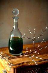 A refined glass decanter filled with dark green liquid, its translucent body reflecting subtle light, stands proudly on a rustic wooden dresser with a smooth, honey-brown finish