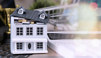 Miniature house filled with money and gold bars, placed on a keyboard with shopping cart and trophy in background, symbolizing financial success.