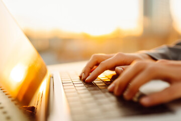 Close-up of hands typing on a laptop in the sunset on a rooftop terrace. A freelancer with a laptop...