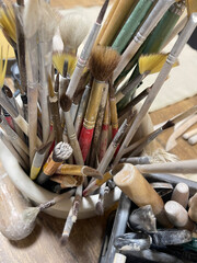 A dense collection of various art brushes (fan, round, flat) with wooden and metal handles, stored in a white container in a messy, creative art studio environment