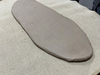a smoothed, flat slab of beige/gray modeling clay resting on a light, textured burlap fabric, representing the raw material for pottery, ceramics, and sculpting arts