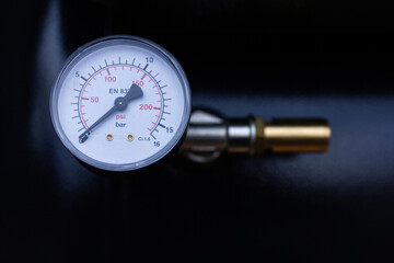 Close-up of a pressure gauge (manometer) for compressed air or gas, displaying dual scales in PSI and Bar, mounted on a metallic connection piece against a dark, blurred background.