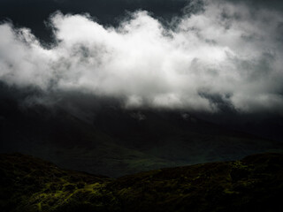 Dark and looming clouds cover the mountain range. Vegetation covers the foreground, while mist and shadows fill the valley during a hike at Coomloughra.