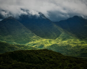 A beautiful view of green mountains shrouded in clouds during a hike along the Coomloughra Horseshoe trail. Light shines on the lower valleys creating a stunning contrast.