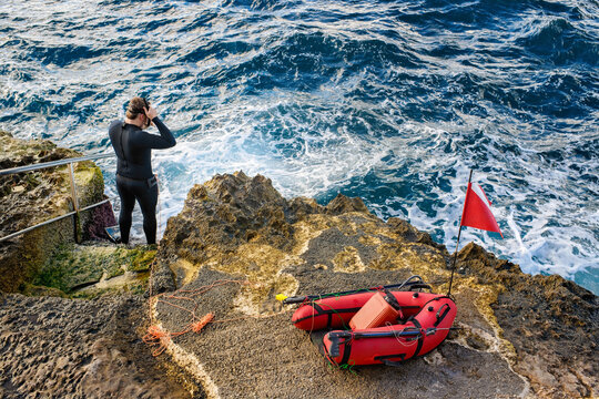 A spearfisherman in a wetsuit prepares to enter the rough, foamy sea from a rocky shore, next to a red buoy and diver's flag. - Powered by Adobe