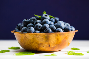 Fresh Blueberries with mint leaves in a wooden bowl on a white wooden table close-up. 