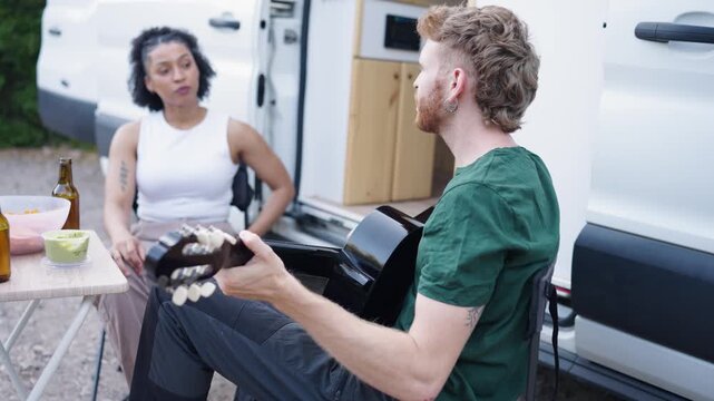 Young man with a mullet playing an acoustic guitar and singing for his girlfriend. Happy diverse couple enjoying a picnic on a van trip