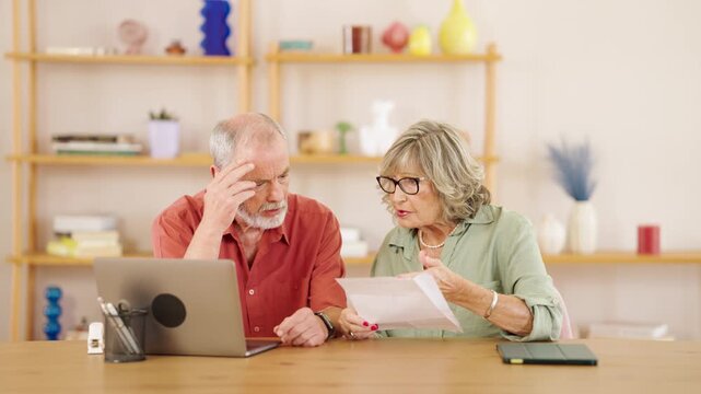 Senior couple analyzing financial documents with laptop, showing concern and discussing potential retirement challenges while sitting together at home