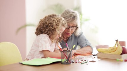 Affectionate grandmother and curly-haired granddaughter spending quality time together, drawing with colorful markers at a wooden table at home
